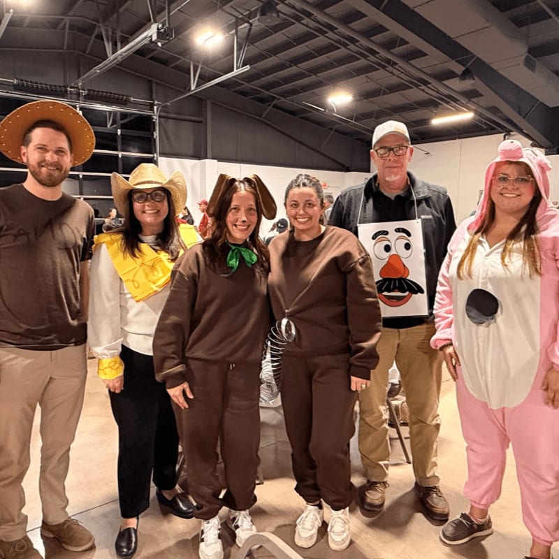 Six people in costumes, including a cowboy, a dog, and a pig, pose in front of a large, open indoor space with a high ceiling.