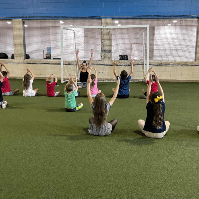 Children in a yoga class sit cross-legged on a green turf floor with a soccer goal in the background.