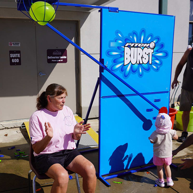 A woman in a pink shirt claps her hands while a toddler in a pink bonnet stands before a blue "Pitch Burst" game, with a green basketball in a hoop overhead.