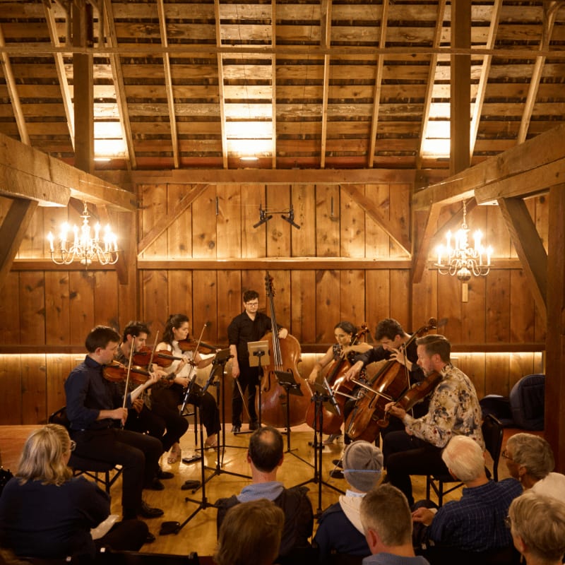 Musicians play string instruments on a wooden stage in a rustic barn, while an audience watches from the foreground.