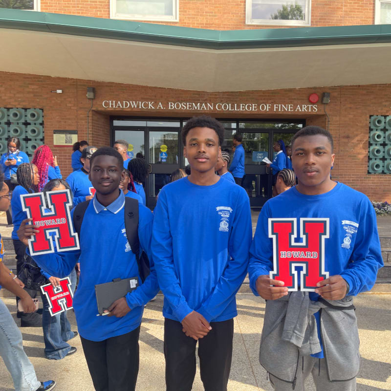 Three young men in blue shirts stand in the foreground holding red "H" signs, with a building labeled "CHADWICK A. BOSEMAN COLLEGE OF FINE ARTS" in the background.