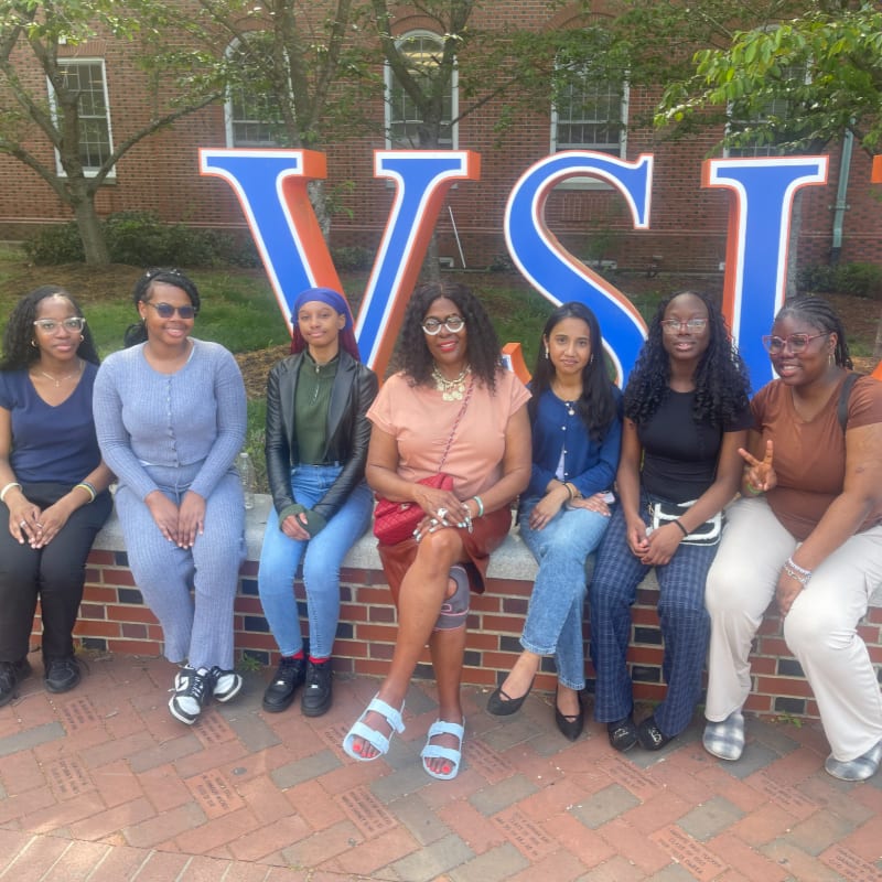 Seven young women pose in front of large blue and orange "VSU" letters, with a brick wall and trees in the background.