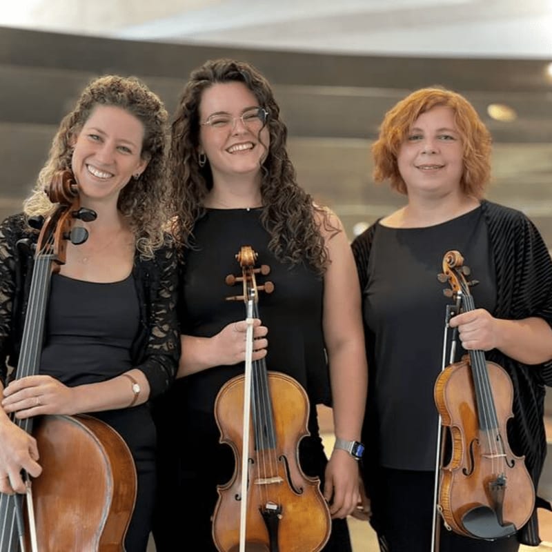 Three smiling women in black attire stand with their string instruments, a cello and two violins, against a blurred, modern architectural background.