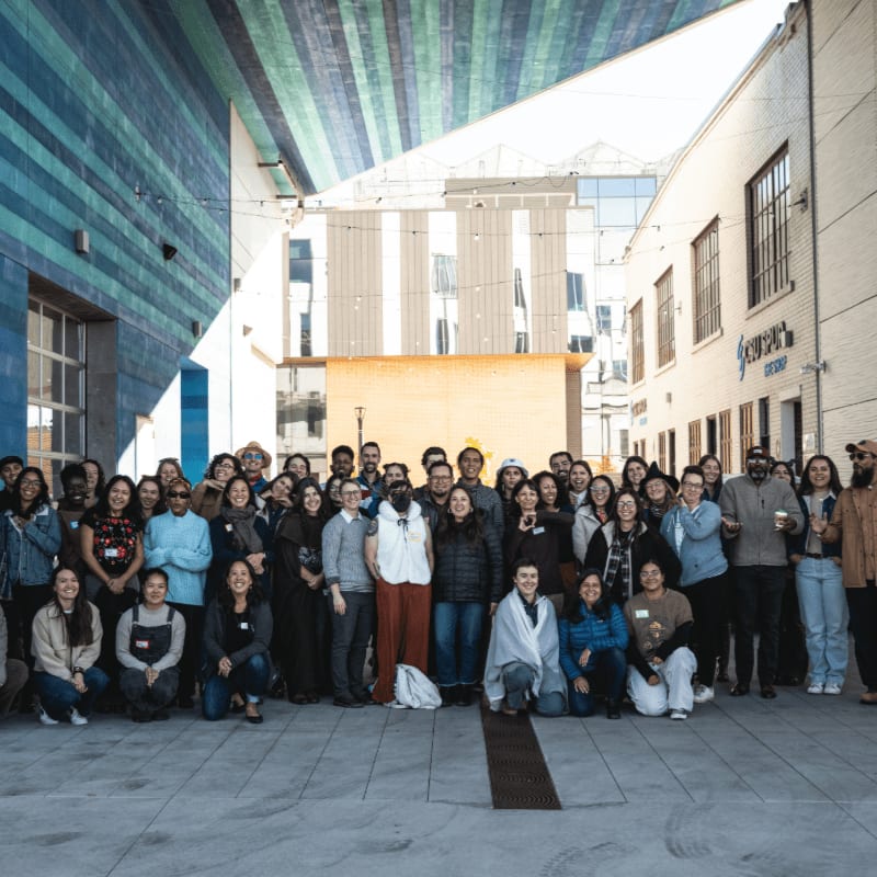 A large group of people pose for a photo in an urban alleyway with colorful buildings in the background.