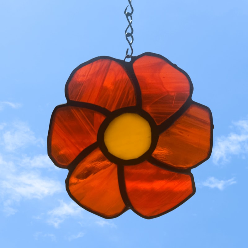 A stained glass flower with orange petals and a yellow center hangs against a bright blue sky with wispy clouds.