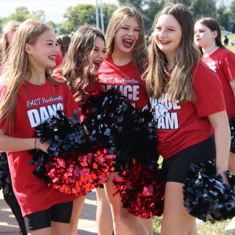 Four smiling girls in red "FACT Pantherettes Dance Team" shirts and black shorts hold red and black pom-poms in the foreground, with other girls and a blurred background of trees and a field.