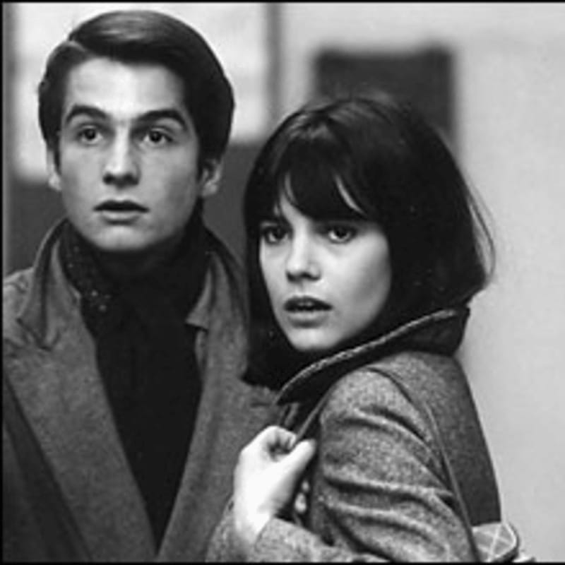 A young man and woman in coats look off to the side with a blurred background.