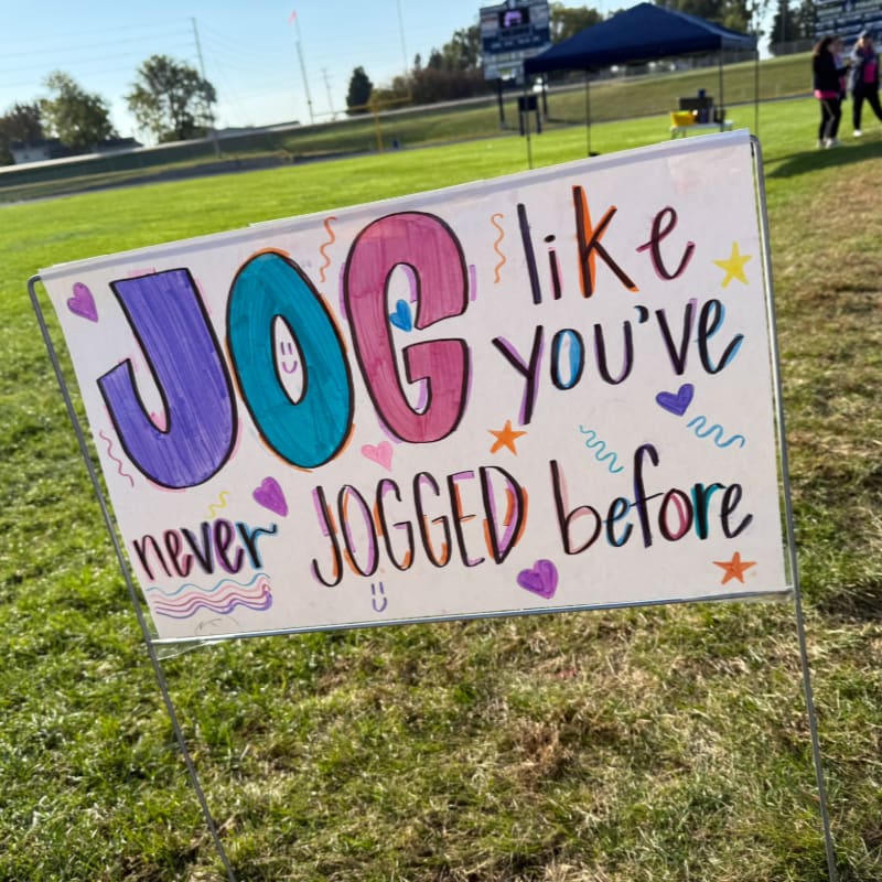 A colorful sign in the foreground encourages people to "JOG like you've never JOGGED before," with a grassy field and a distant tent and spectators in the background.