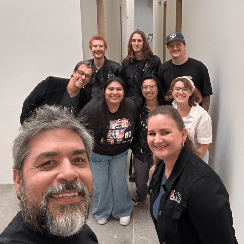 A group of eight smiling people pose for a selfie in a brightly lit hallway with white walls.