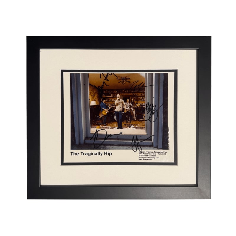 A framed photograph of The Tragically Hip band members performing indoors is in the foreground, with a white mat and black frame surrounding it.