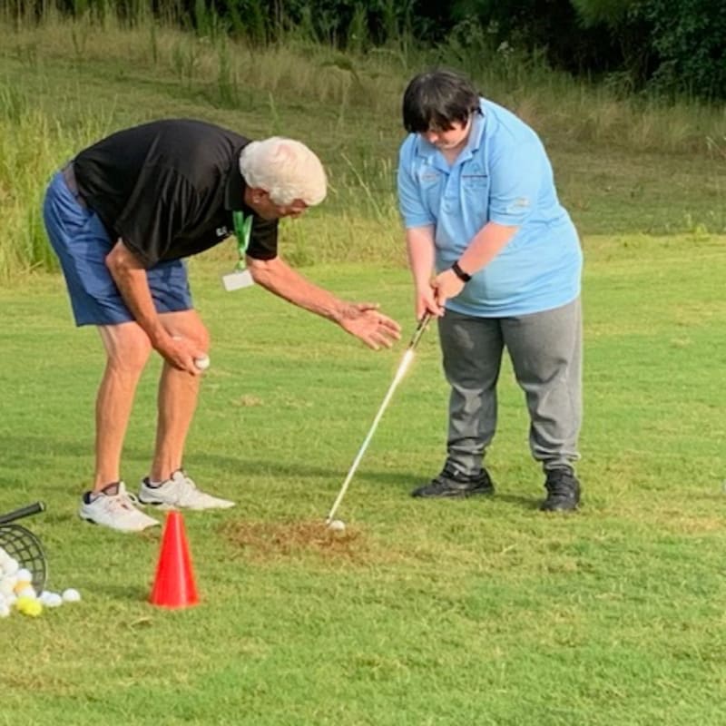 An older man in a black shirt and blue shorts instructs a younger person in a blue shirt and gray pants on a golf course, with a bucket of golf balls and an orange cone in the foreground and a grassy hill with trees in the background.