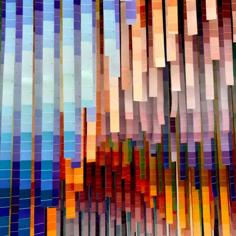 Colorful paint swatches hang in vertical strips, creating a vibrant, abstract pattern against a softly lit background.