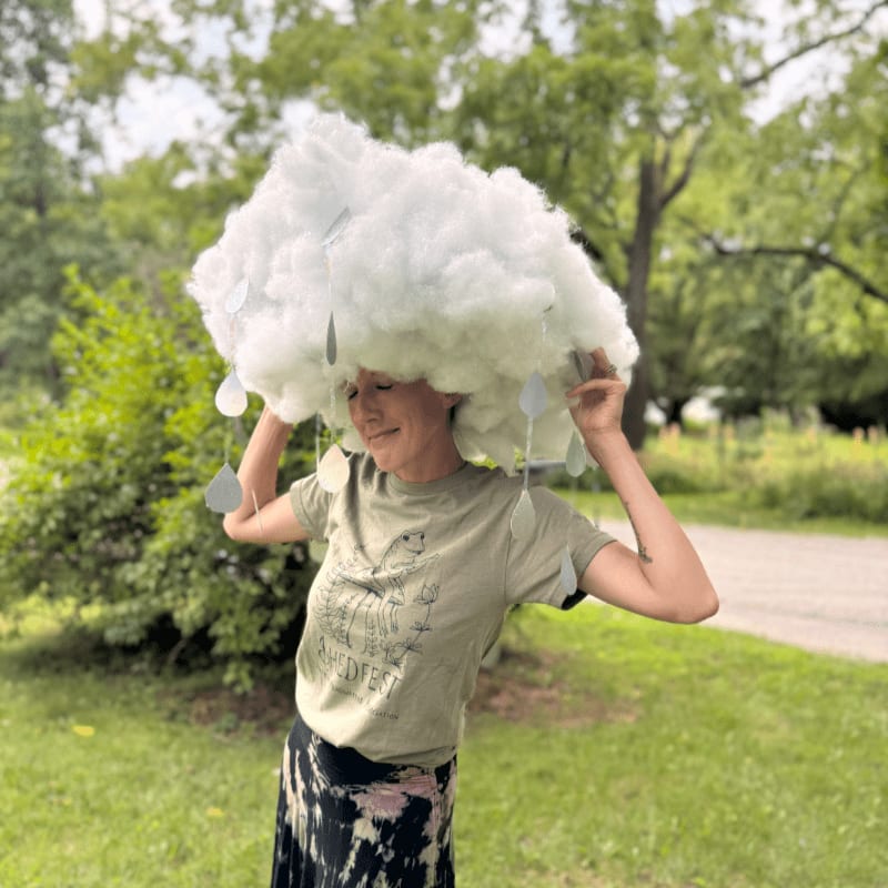 A person in a green t-shirt and patterned skirt wears a large, fluffy cloud costume with dangling raindrops, standing outdoors with lush green trees and a path in the background.