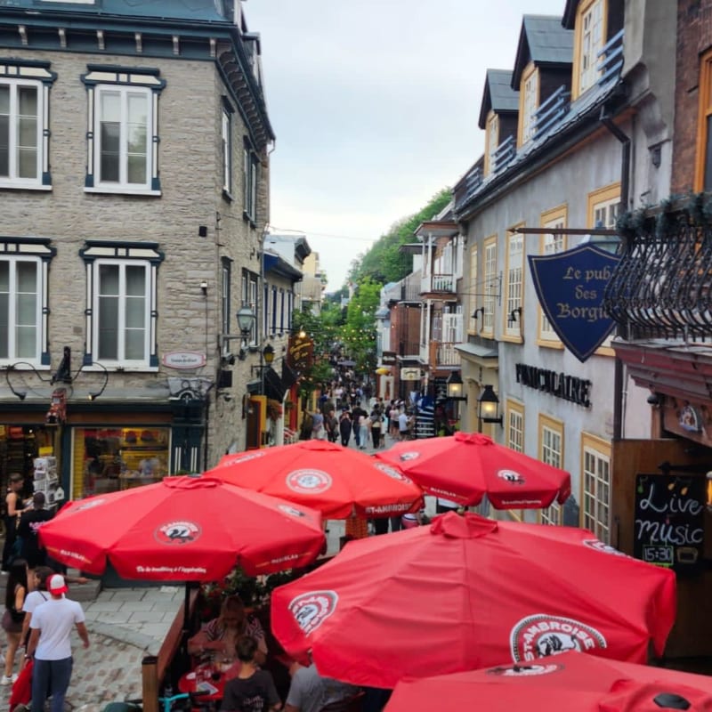 Red umbrellas in the foreground shade outdoor seating on a cobblestone street lined with historic buildings and a crowd of people in the background.