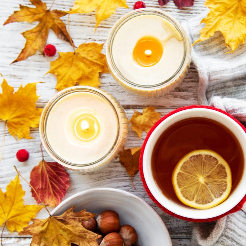 Two lit candles and a cup of tea with a lemon slice sit amongst scattered autumn leaves and hazelnuts on a wooden surface.