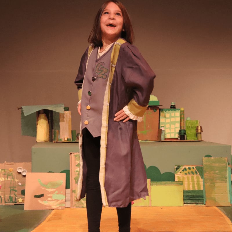 A young girl in a costume stands on a yellow stage in front of a backdrop of colorful, cartoonish buildings.