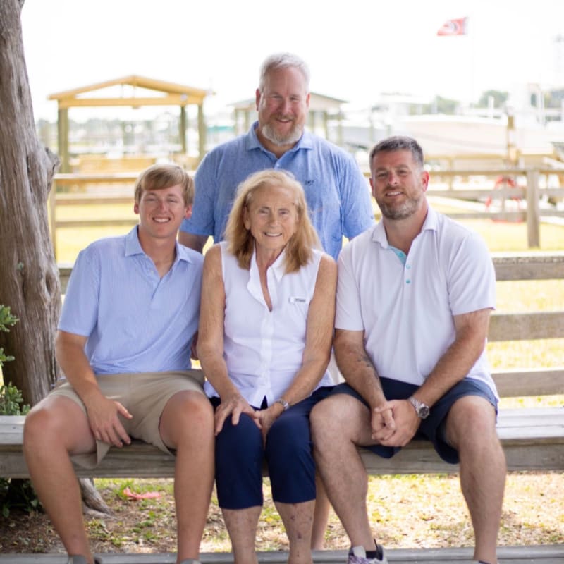 A family of four poses for a photo on a bench in the foreground, with a waterfront scene featuring boats and buildings in the background.