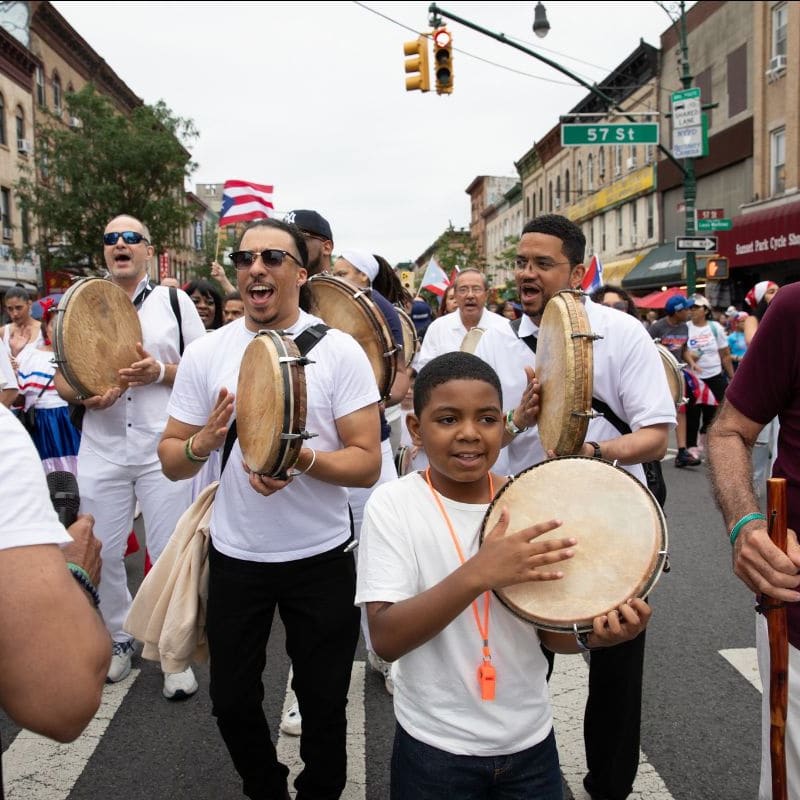 A group of people in white shirts play drums in the foreground while a street with buildings and a traffic light forms the background.