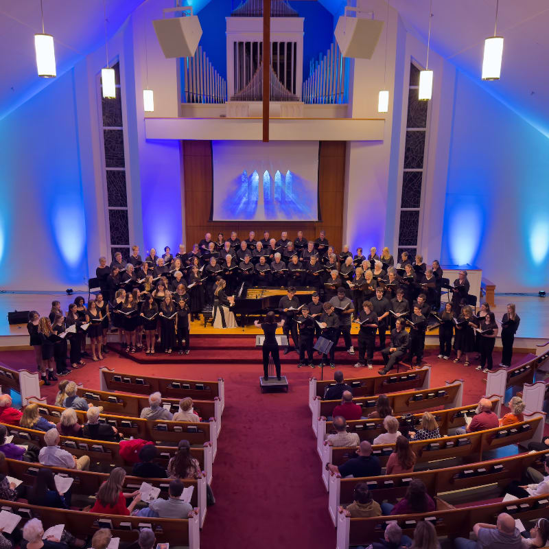 A choir performs on a stage in front of an organ and a screen, while an audience watches from pews in the foreground.
