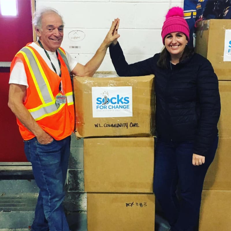 A man in a safety vest and a woman in a pink hat give each other a high-five over boxes of socks for change, with more boxes stacked in the background.