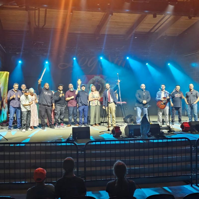 A group of people stand on a stage with bright blue spotlights in the background, while a few audience members are visible in the foreground.