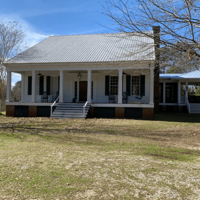 A white farmhouse with a wraparound porch sits on a grassy lawn under a bright blue sky.