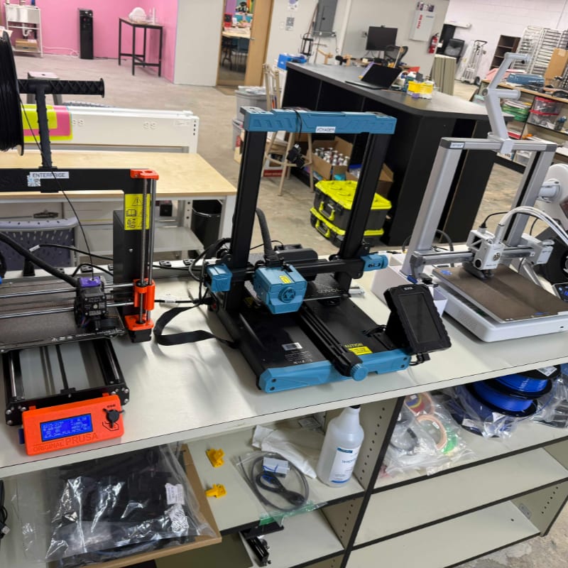 Three 3D printers sit on a workbench in the foreground, with a cluttered workshop visible in the background.