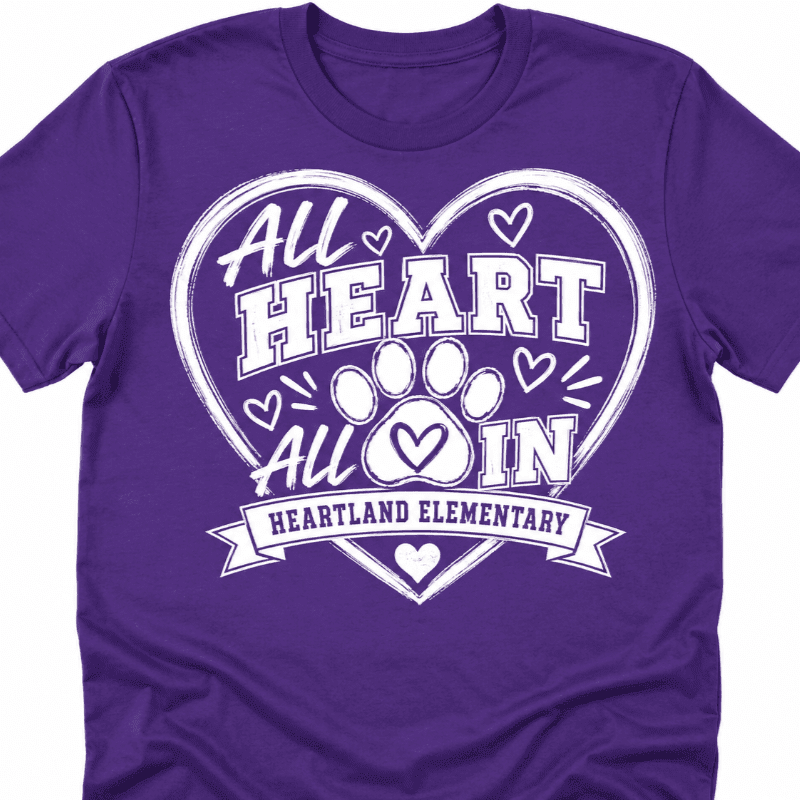 A purple t-shirt features a white heart graphic with paw prints and the text "ALL HEART ALL IN HEARTLAND ELEMENTARY" in the foreground, against a plain white background.