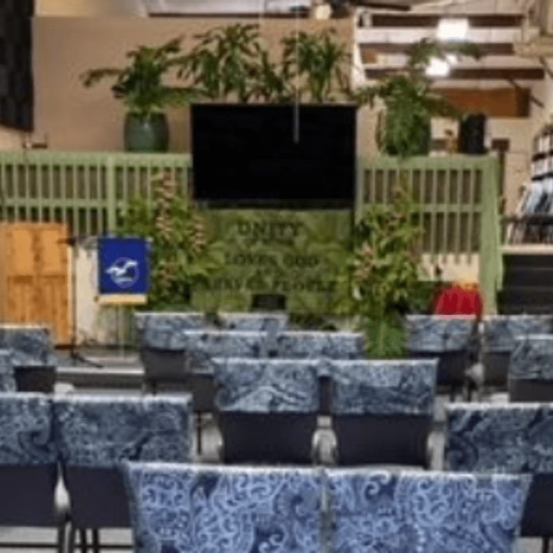 Rows of chairs with patterned covers fill the foreground, leading to a stage with a large screen, plants, and a banner that reads "UNITY LOVES GOD AND HIS PEOPLE" in the background.