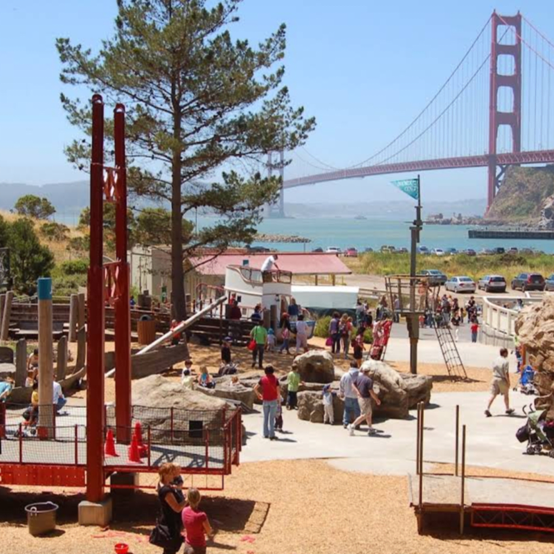 A vibrant playground with children and adults enjoying themselves in the foreground is set against the iconic Golden Gate Bridge and a sparkling bay in the background.