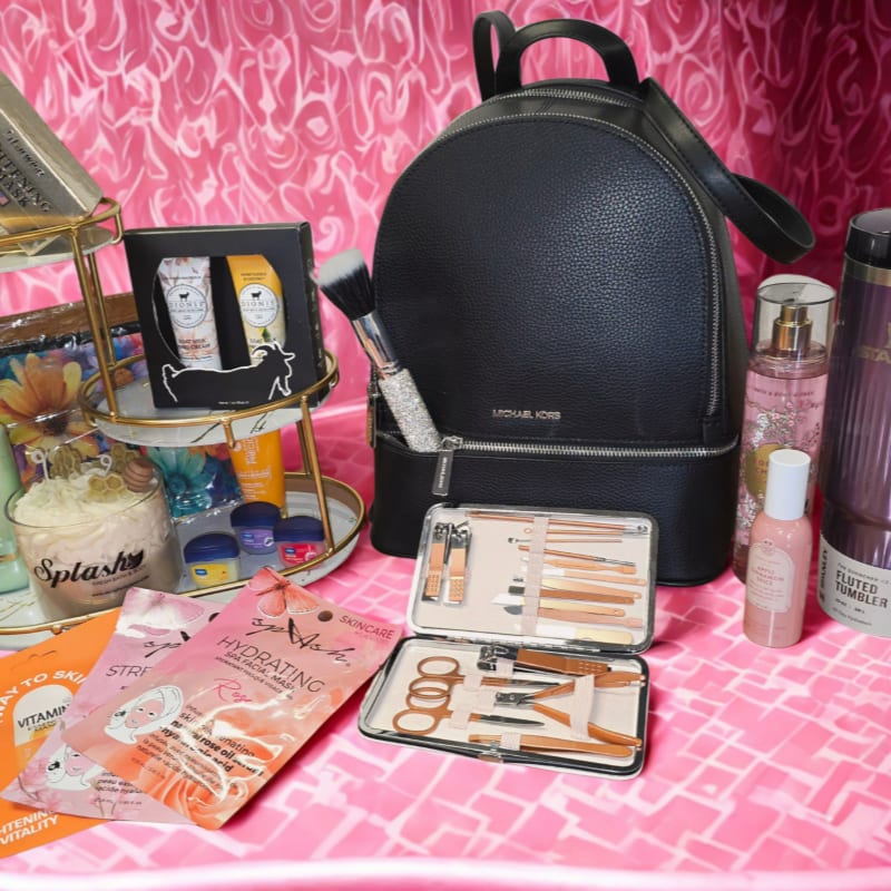 A black Michael Kors backpack sits behind a collection of skincare products and makeup tools, all arranged on a pink, textured surface.