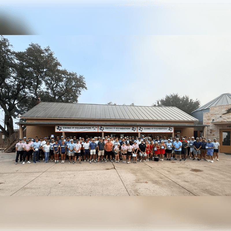 A large group of people stands in front of a building with a banner that reads "SWING IT FOUNDATION," while trees frame the background.