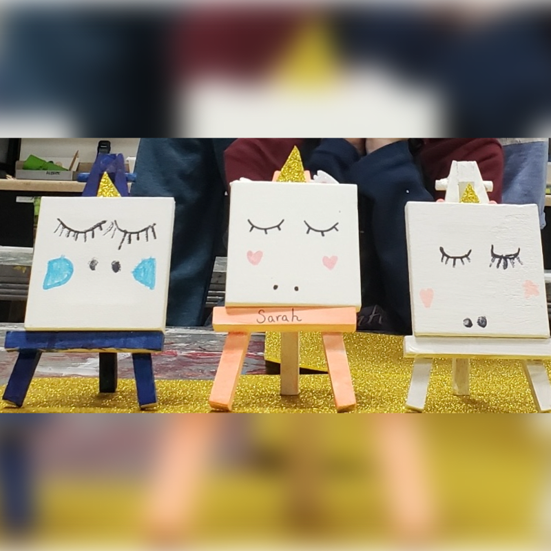 Three small, easel-mounted paintings of unicorn faces with closed eyes and rosy cheeks sit on a glittery surface, with blurred figures in the background.