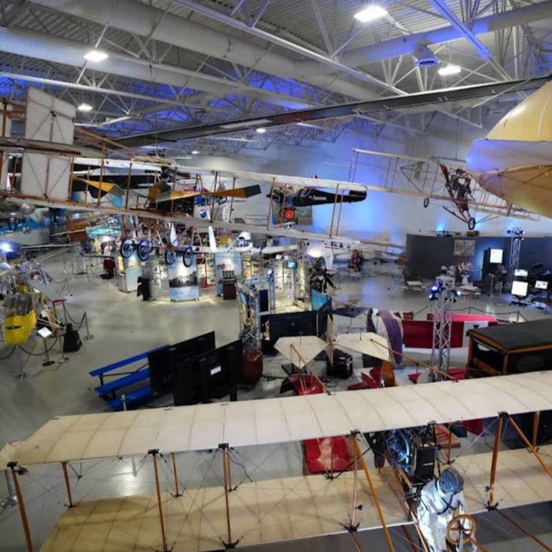 In the foreground, a vintage biplane with a fabric wing is displayed, while the background reveals a large hangar filled with various aircraft and exhibits.