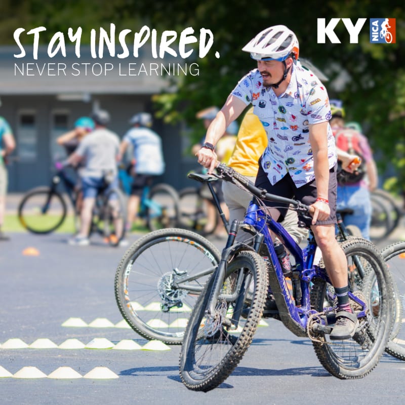 A man in a colorful shirt and helmet rides a purple mountain bike in the foreground, while other cyclists and a building are blurred in the background.