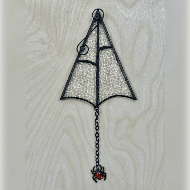A stained glass spiderweb ornament with a dangling spider hangs against a light wood grain background.