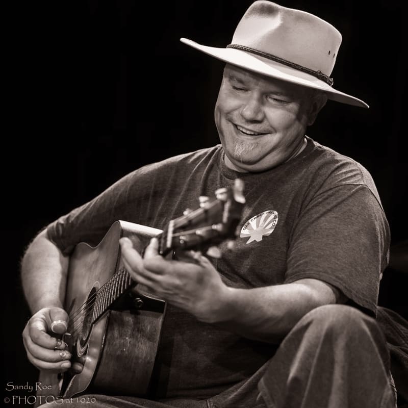 A man in a hat smiles while playing a guitar against a dark background.