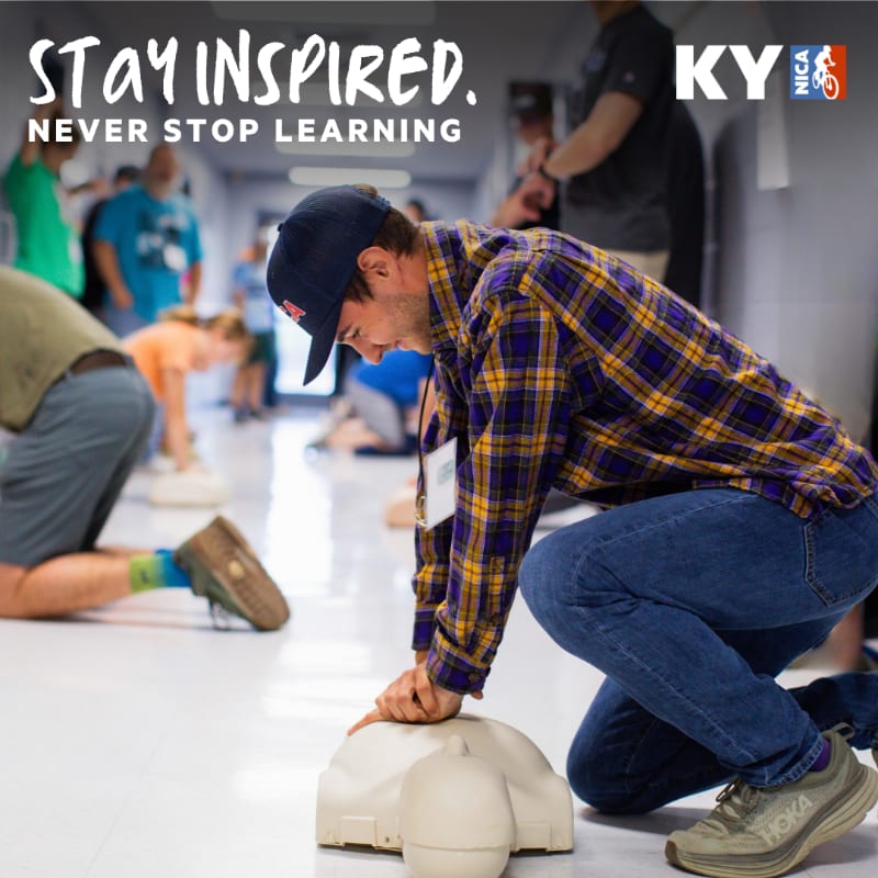 A man in a plaid shirt practices CPR on a dummy in the foreground, while others in a hallway with "Stay Inspired. Never Stop Learning." text and a NICA logo are in the background.