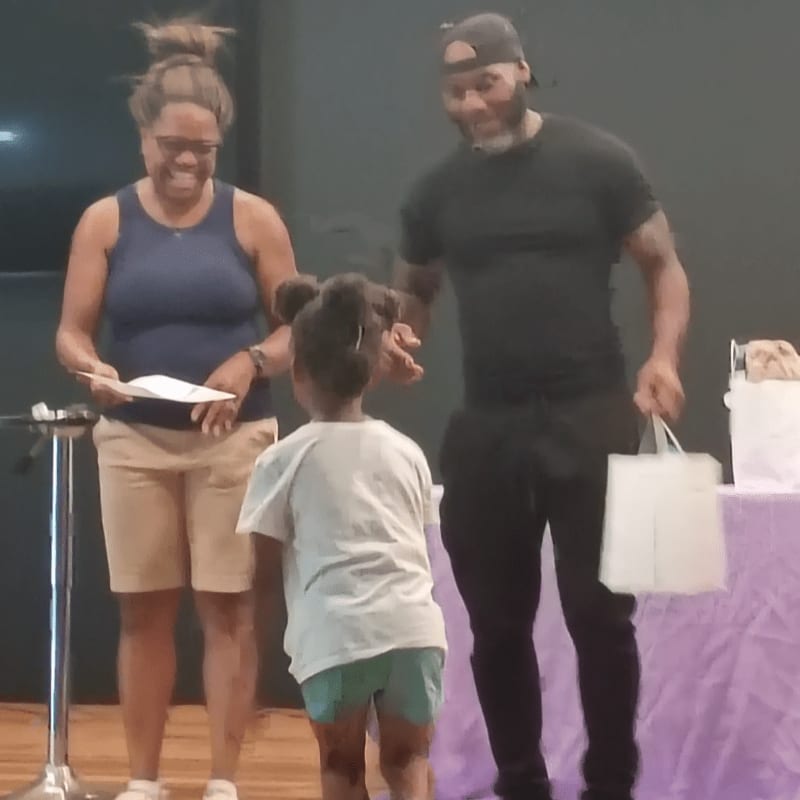 A woman and a man stand on a stage with a young girl in the foreground, while a plain dark background is visible behind them.