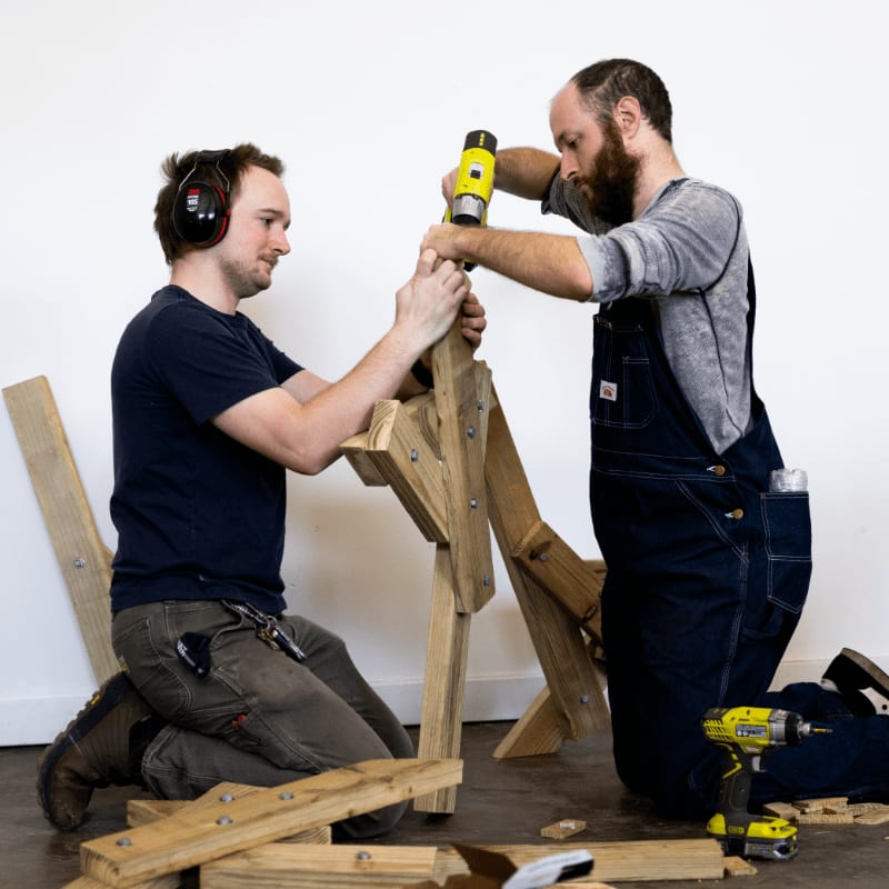 Two men in overalls and work clothes kneel on a wooden floor, assembling a wooden structure with power tools against a plain white background.