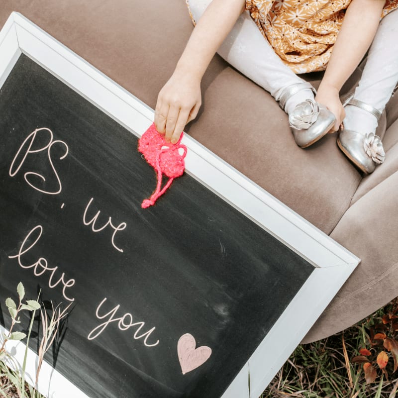 A child's hand erases "PS. we love you" from a chalkboard with a pink heart, while sitting on a brown couch outdoors.