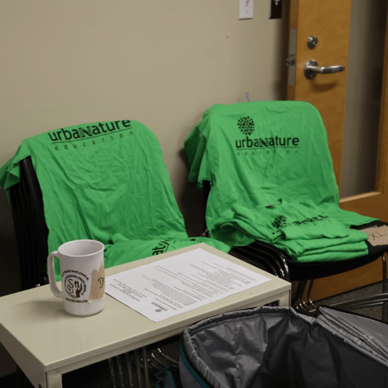 Two green t-shirts with "urban NATURE education" printed on them are draped over chairs in the foreground, with a table holding a mug and papers in front of them, while a closed door is visible in the background.