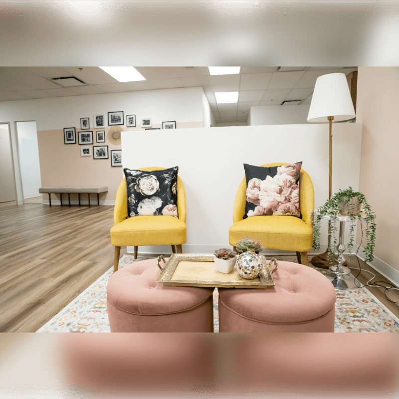 Two plush pink ottomans with a decorative tray sit in the foreground before two bright yellow chairs adorned with floral pillows, while a gallery wall of framed photos hangs on a pale pink wall in the background.