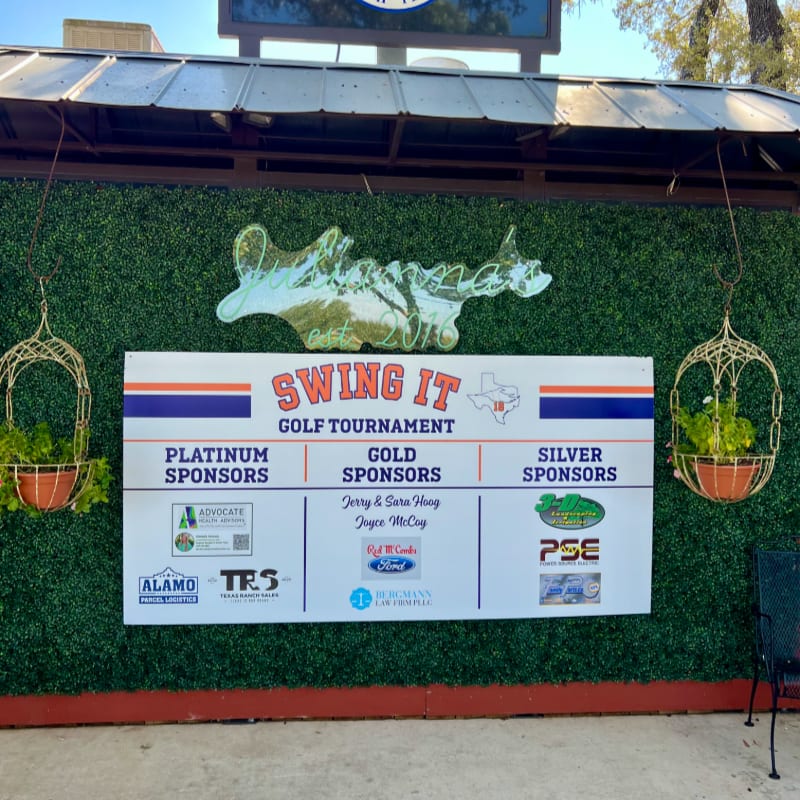 A green hedge wall serves as the backdrop for a "Swing It Golf Tournament" banner, with hanging planters on either side and a metal airplane decoration above.