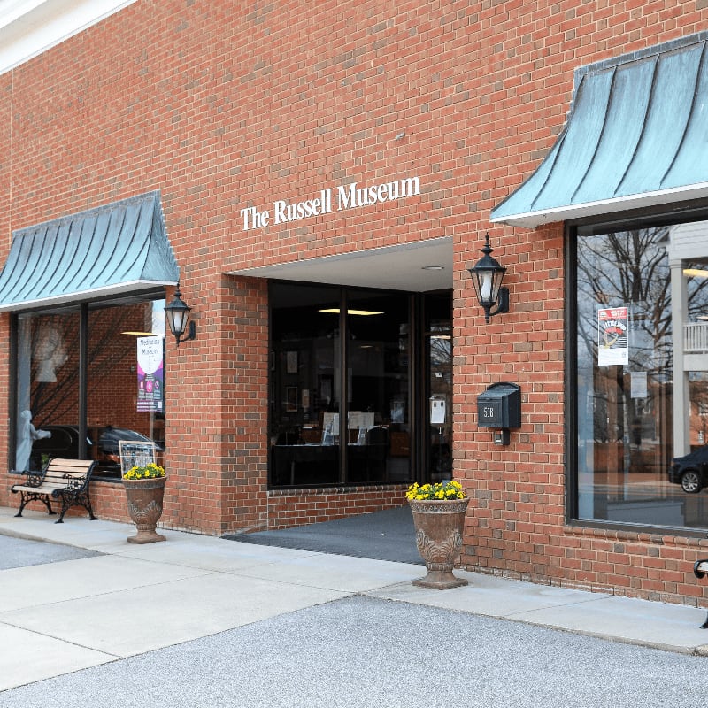Two ornate planters flank the entrance to The Russell Museum, a brick building with large display windows and awnings.