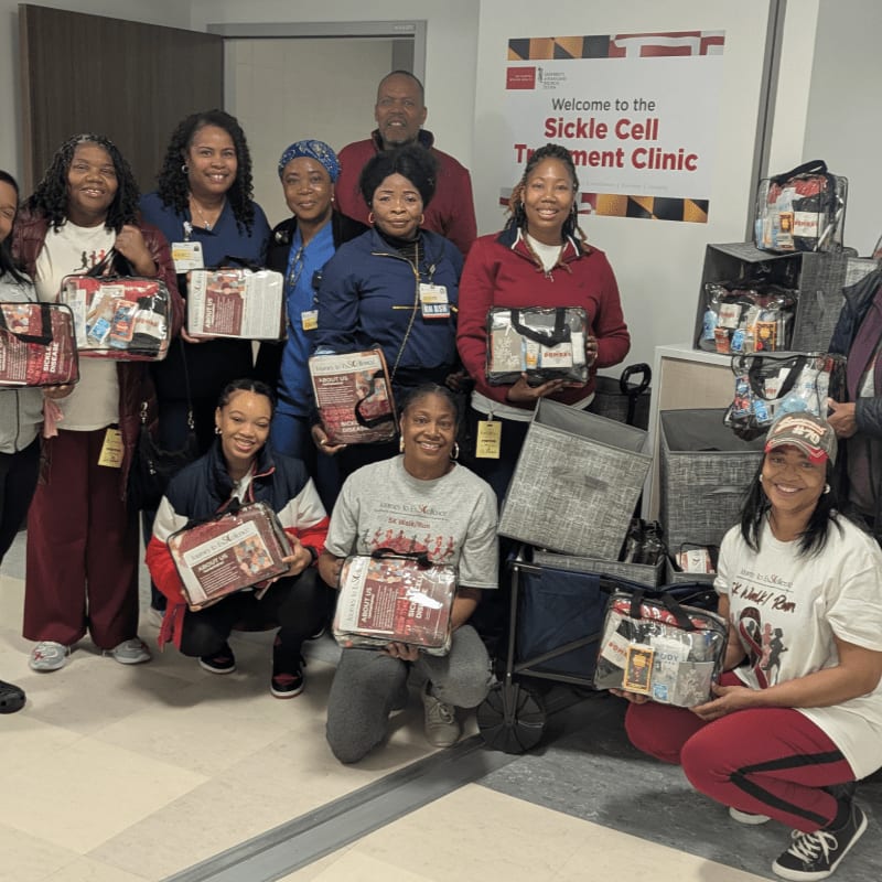 A group of people stand in a clinic hallway, holding gift bags and blankets, with a sign welcoming visitors to the Sickle Cell Treatment Clinic in the background.