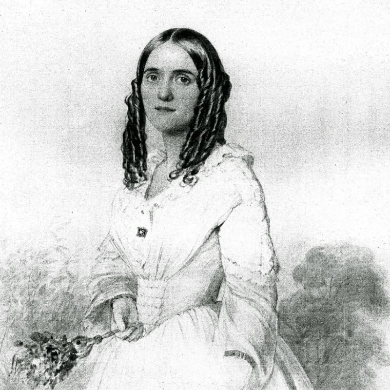A woman in a white dress with dark curly hair holds a bouquet of flowers in the foreground, set against a softly rendered background of foliage.