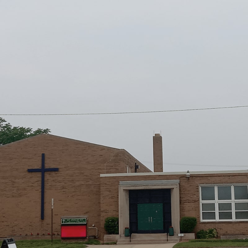 A brick church with a large blue cross on its side stands under a cloudy sky, with a green double door entrance in the foreground.