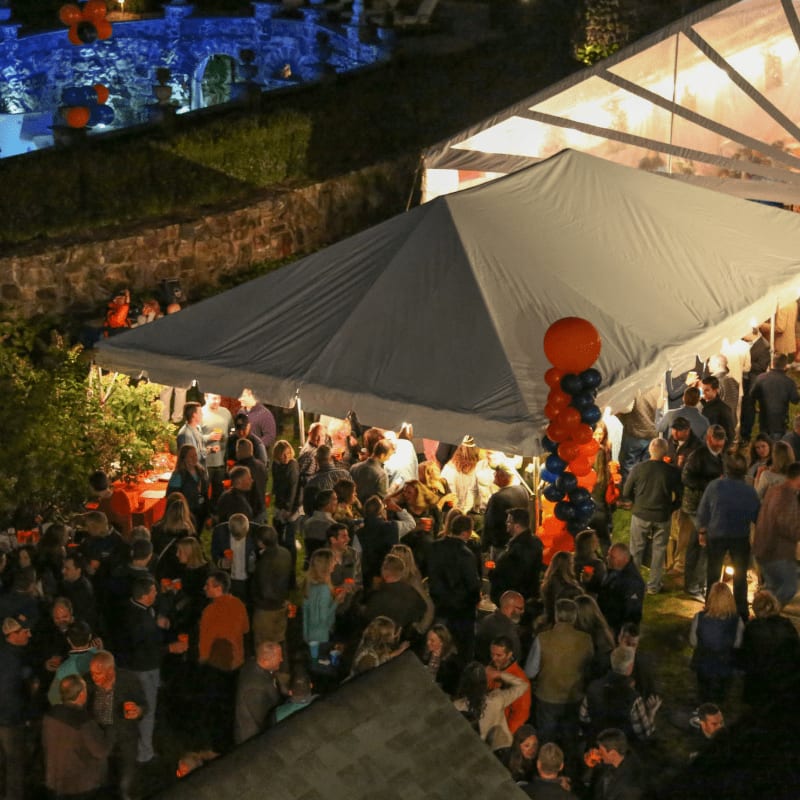A large crowd of people are gathered under a white tent in the foreground, while a pool and illuminated stone structures are visible in the background.