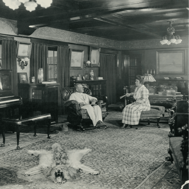 A man and woman converse in a richly decorated room with a lion skin rug in the foreground and a piano and bookshelves in the background.